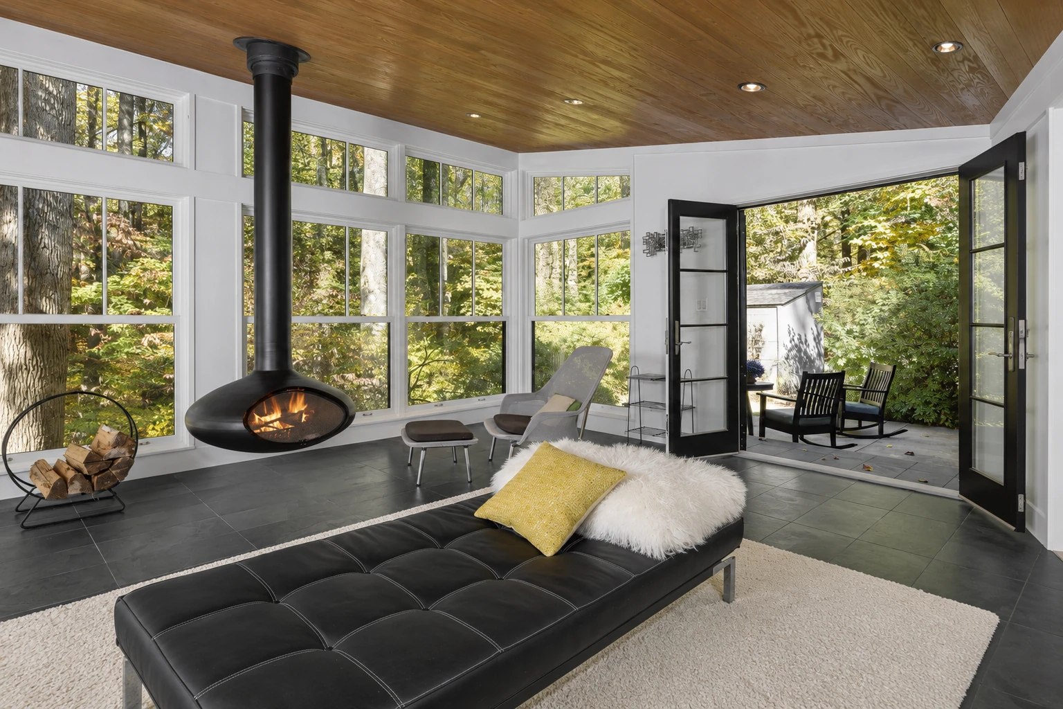 Bright four-season sunroom with broad windows, a wood ceiling, a lit suspended fireplace and open doors to a patio.