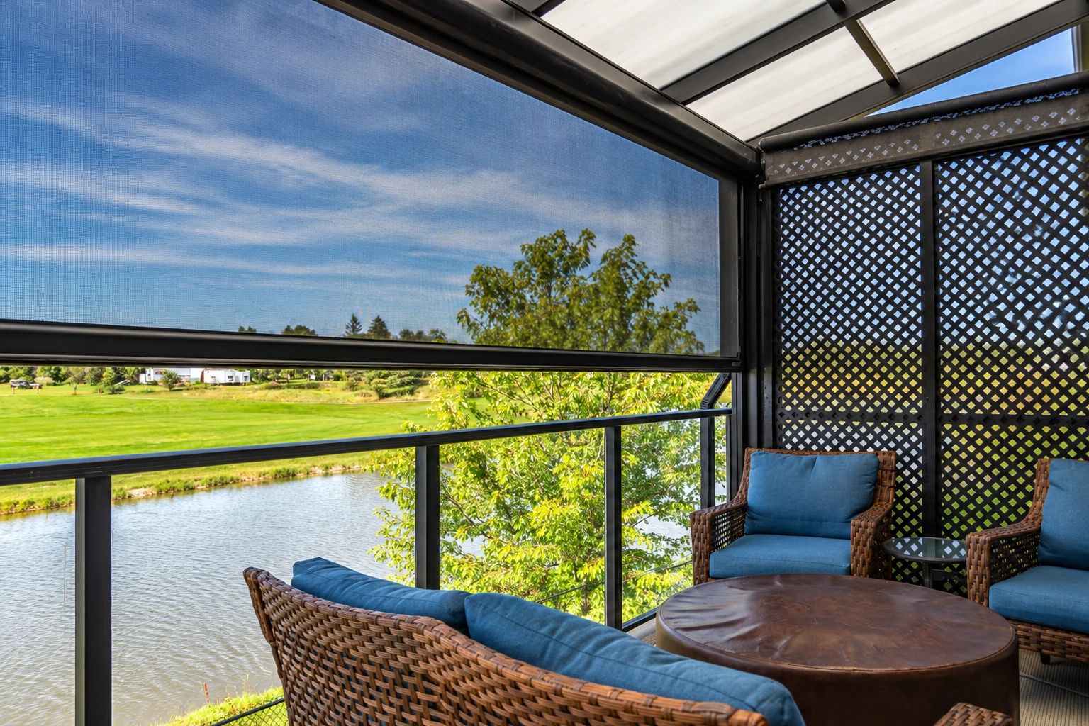 A covered patio with a mesh shade, lattice panels, and wicker seating overlooks a green lawn and water.