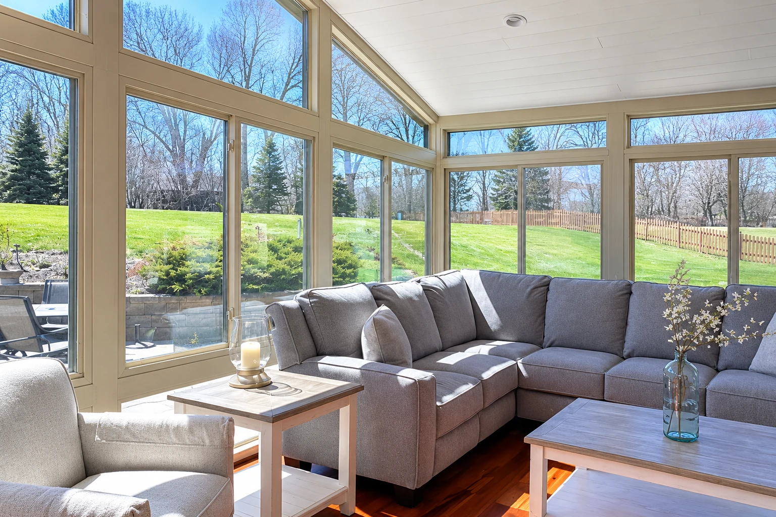 A bright sunroom with large beige-framed windows and a gray sectional sofa looks out onto a green lawn.