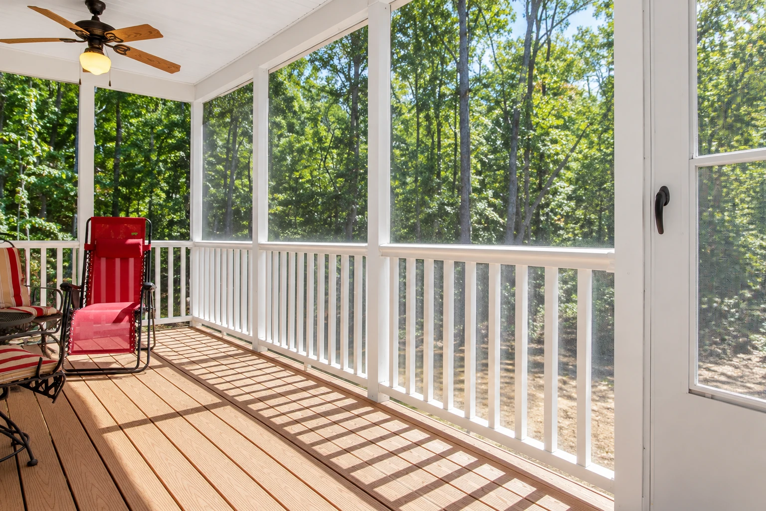 A screened porch with white framing and a wood-plank floor overlooks leafy trees; red chairs sit beneath a ceiling fan.
