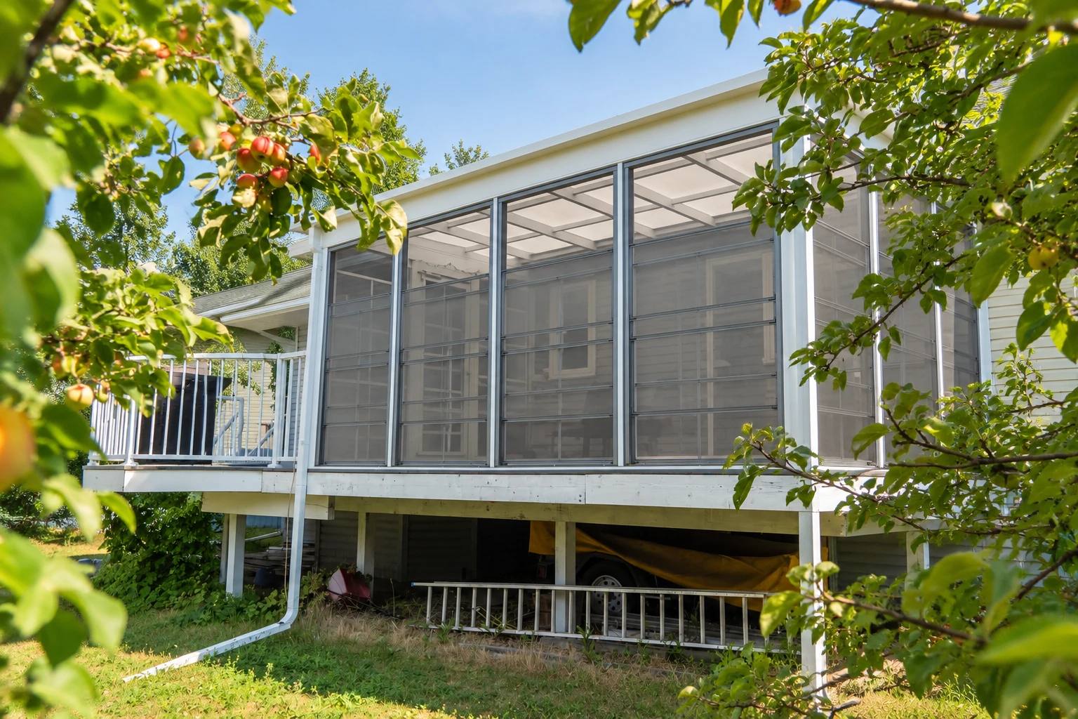 Raised screened sunroom addition with white framing and a sloped roof, partly shaded by leafy fruit trees.