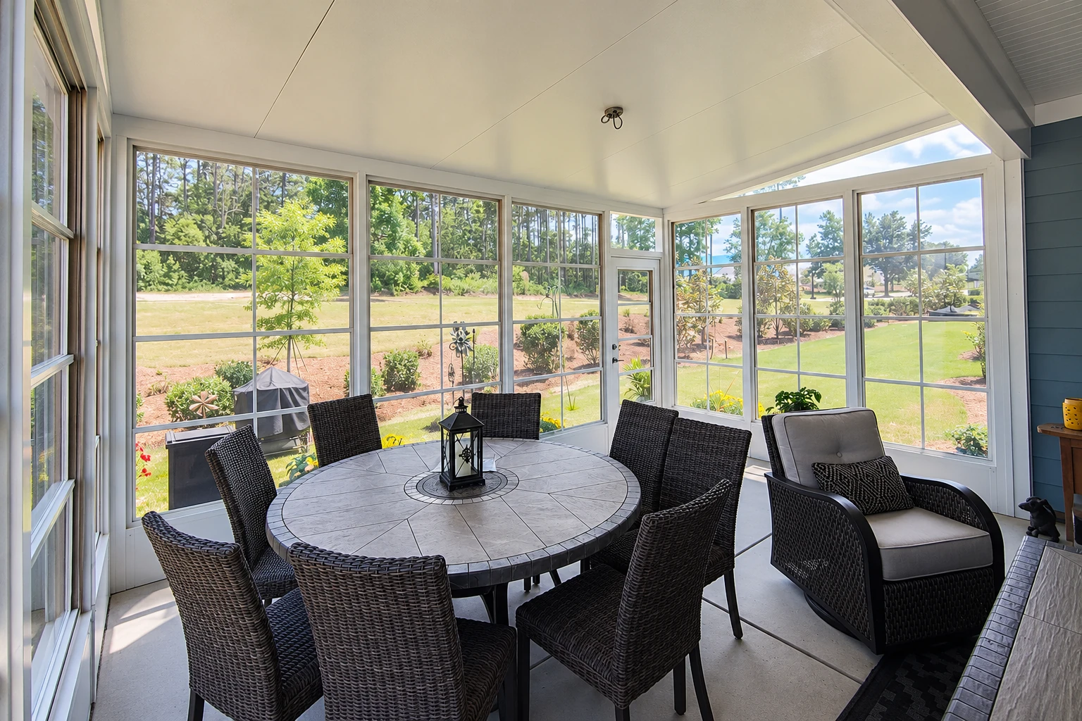 Enclosed patio sunroom with white-framed windows, a round dining table and dark wicker chairs overlooking a lawn.