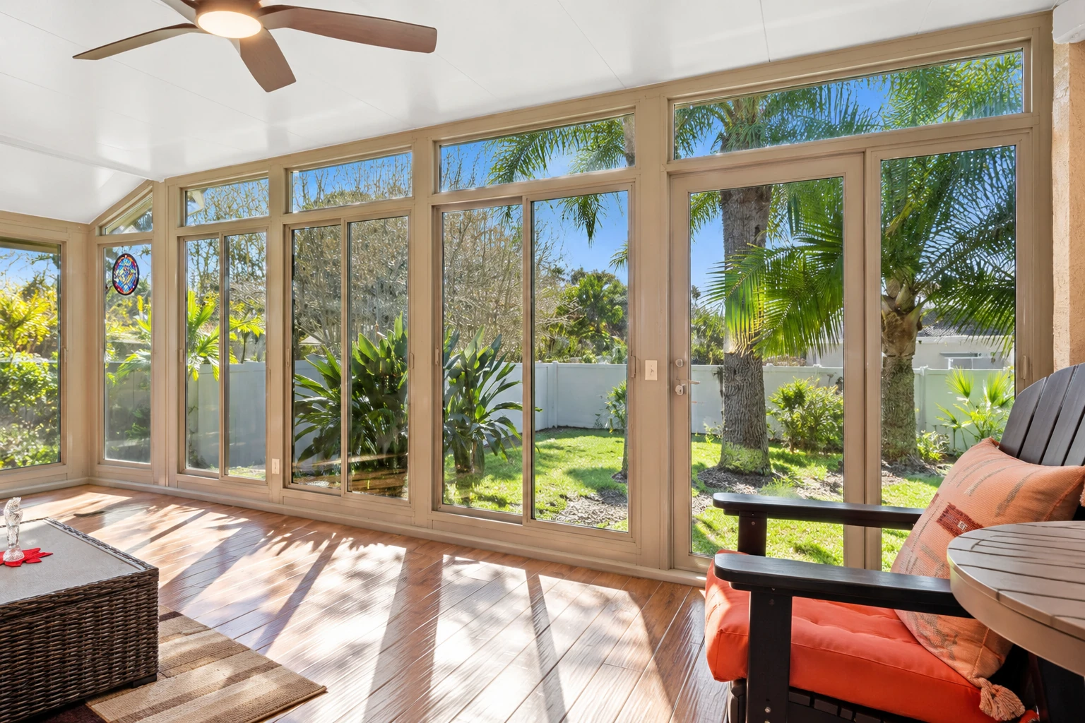 Sunroom interior with tan-framed windows, a ceiling fan and orange-cushioned chairs overlooking a garden.
