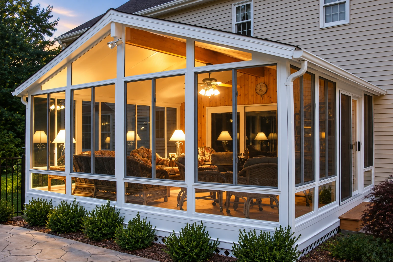 White-framed vinyl sunroom attached to a house, with a peaked glass front and warmly lit wicker seating inside.